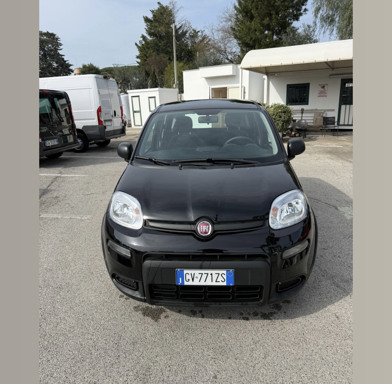 Fiat Panda nera vista di fronte, targa CV771ZS, in un parcheggio con furgoni bianchi e prefabbricati tra gli alberi