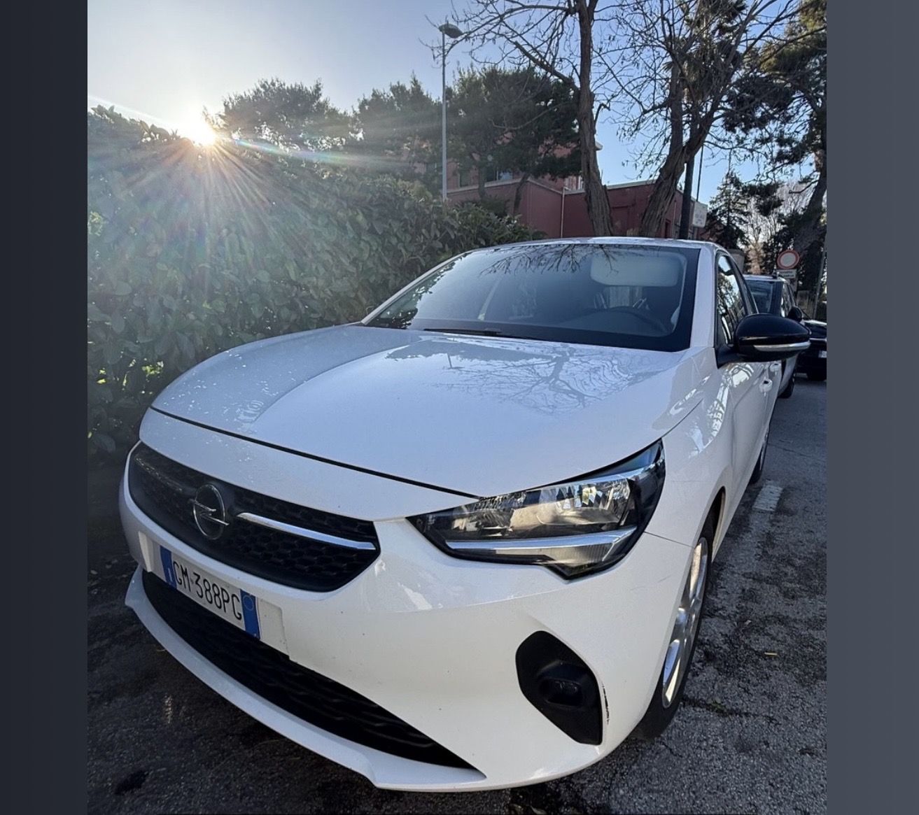 Opel Corsa bianca parcheggiata in strada accanto a una siepe, con alberi spogli, sole basso e un edificio rosso sullo sfondo