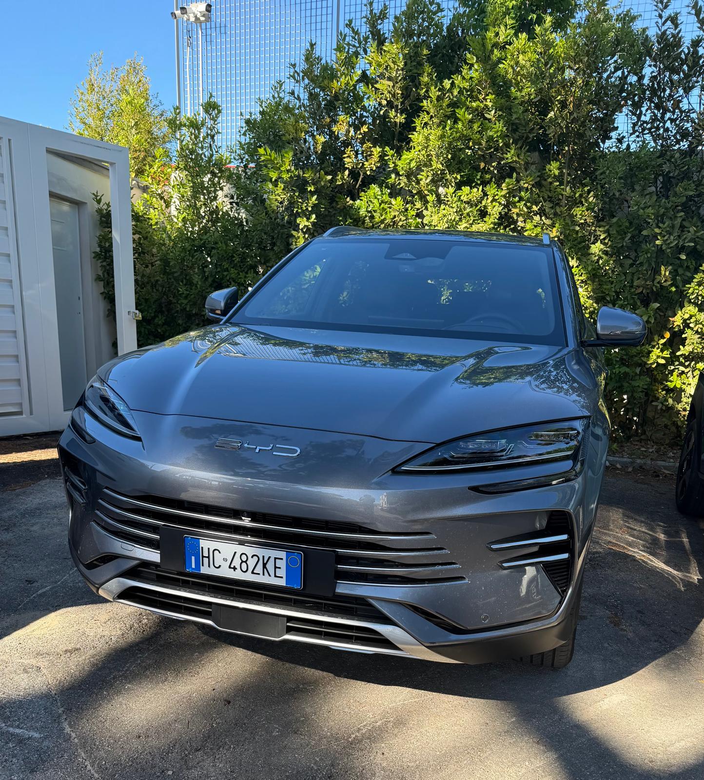 SUV BYD grigio metallizzato con targa HC 482KE parcheggiato su asfalto davanti a una siepe verde e a un box bianco