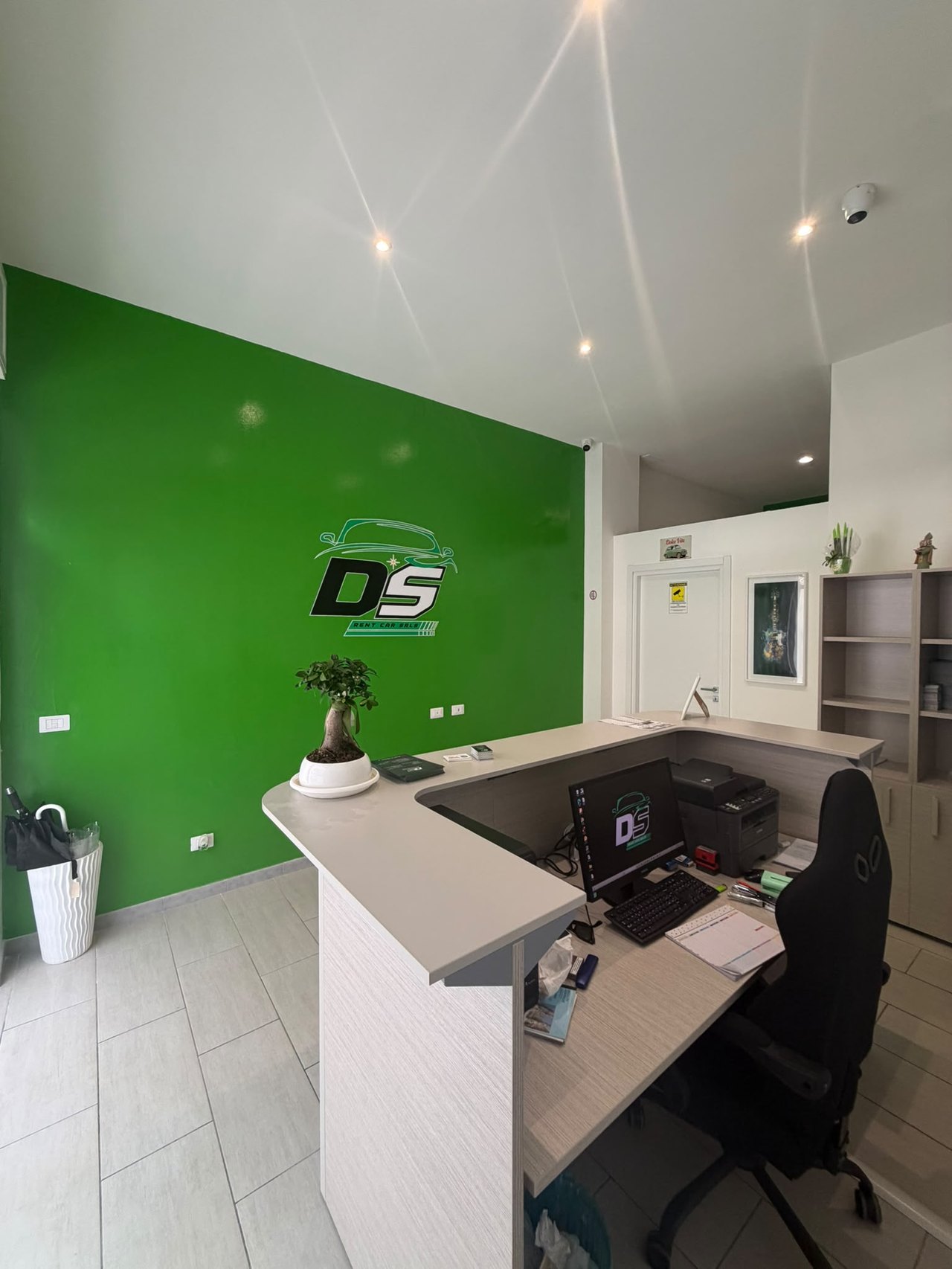 Reception di DS Rentcar con logo DS su parete verde, bancone bianco ad angolo, bonsai, monitor e sedia nera